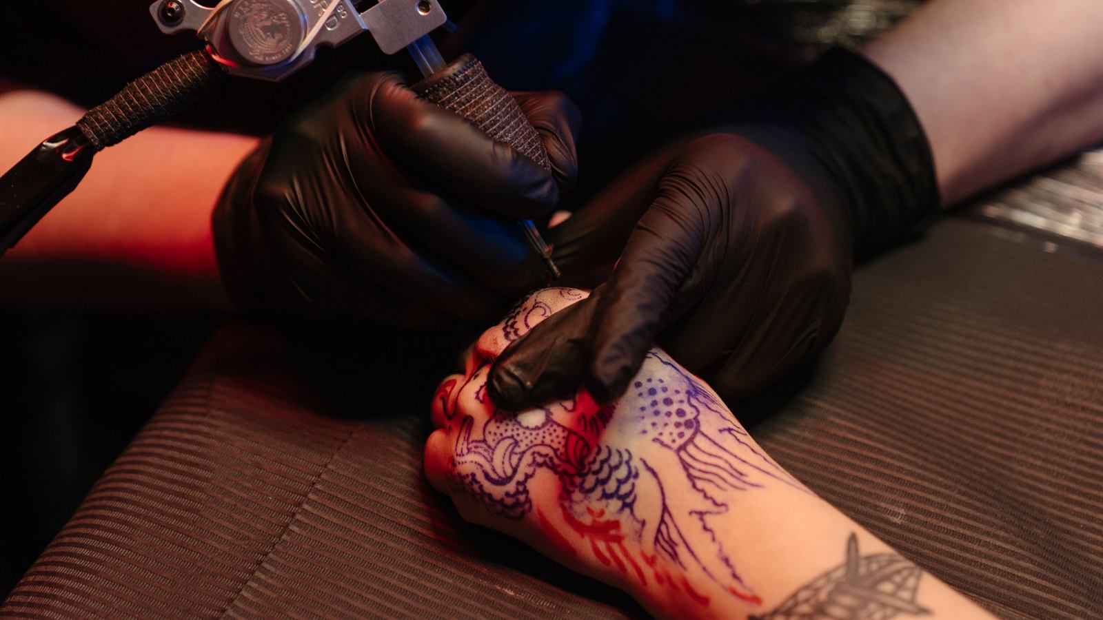 Tattoo artist working on a hand piece under red and blue studio light