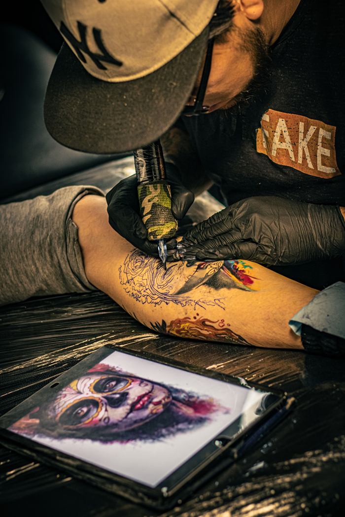 Artist tattooing an ornamental sleeve on a forearm, reference sheet resting nearby